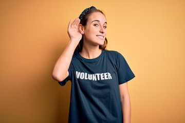 Young beautiful woman wearing volunteer t-shirt doing volunteering over yellow background smiling with hand over ear listening an hearing to rumor or gossip. Deafness concept.