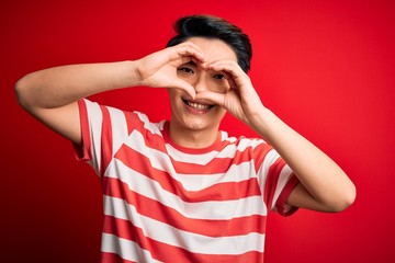 Young handsome chinese man wearing casual striped t-shirt standing over red background Doing heart shape with hand and fingers smiling looking through sign
