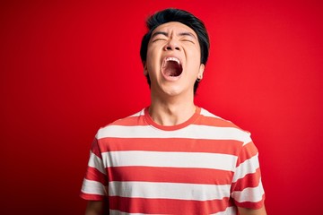 Young handsome chinese man wearing casual striped t-shirt standing over red background angry and mad screaming frustrated and furious, shouting with anger. Rage and aggressive concept.