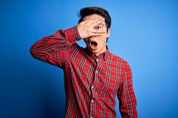 Young handsome chinese man wearing casual shirt and glasses over blue background peeking in shock covering face and eyes with hand, looking through fingers with embarrassed expression.