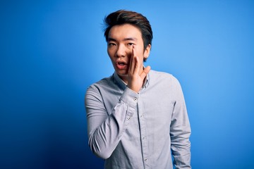 Young handsome chinese man wearing casual shirt standing over isolated blue background hand on mouth telling secret rumor, whispering malicious talk conversation