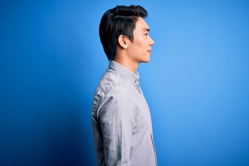 Young handsome chinese man wearing casual shirt standing over isolated blue background looking to side, relax profile pose with natural face and confident smile.