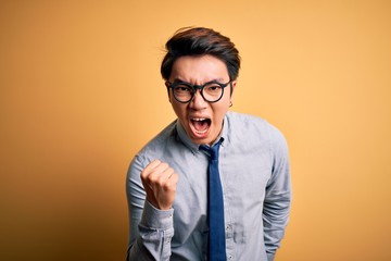 Young handsome chinese businessman wearing glasses and tie over yellow background angry and mad raising fist frustrated and furious while shouting with anger. Rage and aggressive concept.
