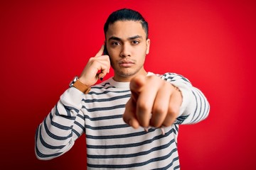Fototapeta premium Brazilian man having conversation talking on the smartphone over isolated red background pointing with finger to the camera and to you, hand sign, positive and confident gesture from the front