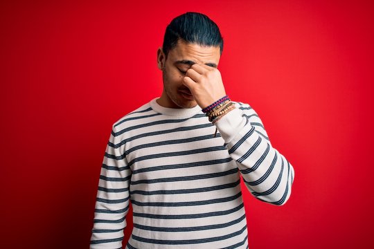 Young brazilian man wearing casual striped t-shirt standing over isolated red background tired rubbing nose and eyes feeling fatigue and headache. Stress and frustration concept.