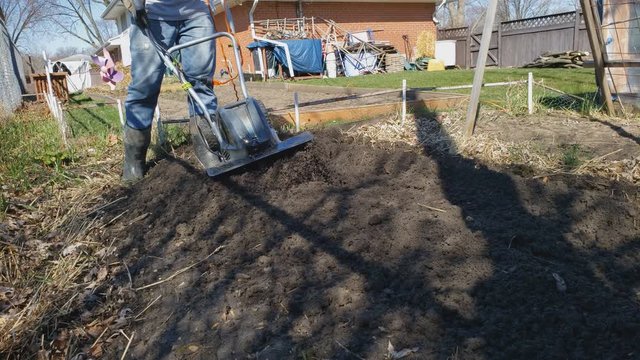 Home gardening - Man in boots using electric soil cultivator or tiller preparing back yard garden soil for spring planting by softening and breaking apart clumps of soil.