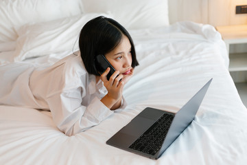 Fototapeta premium Beautiful asian female in white shirt using mobile phone while working on bed. work from home concept.