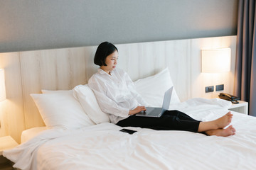 Beautiful asian female in white shirt using laptop while working on bed. work from home concept.