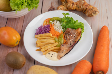 Fish steak with french fries, kiwi, lettuce, carrots, tomatoes, and cabbage in a white dish.