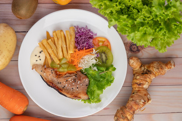 Fish steak with french fries, kiwi, lettuce, carrots, tomatoes, and cabbage in a white dish.