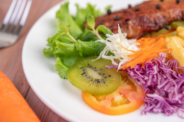 Fish steak with french fries, kiwi, lettuce, carrots, tomatoes, and cabbage in a white dish.