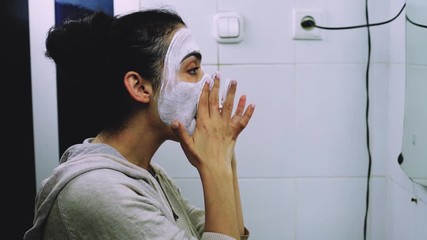 Woman applying facial cosmetic mask in the bathroom. Mixed-race female apply white nourishing mask or creme on face. Applying mask on face. Skincare cleansing spa relax concept