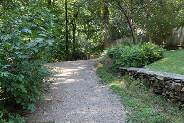 Gravel path leading to a crest in the road, bordered by vegetation, retaining wall, and grass, horizontal aspect
