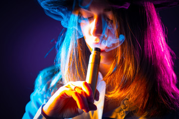 A girl with long hair holds a VAPE in her hands and looks at it. A woman in a cloud of smoke from...