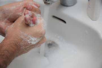 Manos de hombre frontandose con jabon para lavarse y prevenir el coronavirus. grifo metalico y lavabo blanco de fondo. 