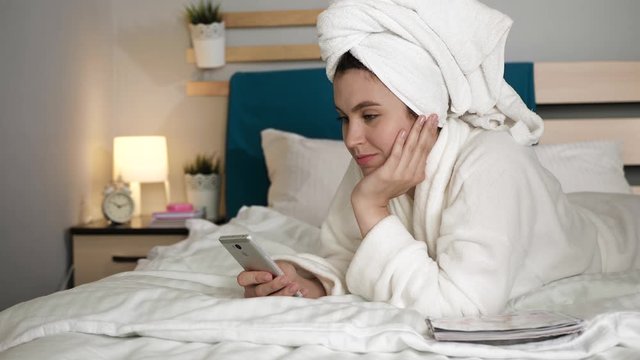 Girl scrolling phone. Woman in bedroom with towel on her head and in white coat lies on stomach on bed and leafs through her cell news feed. Medium shot