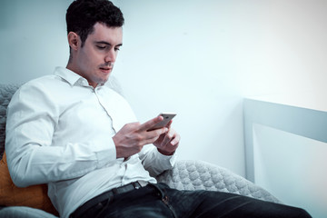 cinematic tone of young Caucasian man using smartphone or cell phone and sitting alone on chair in white room