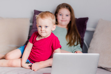 Two children a boy and a girl lie on a sofa in home clothes and watch videos on a laptop and look at camera for a moment.
