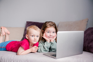 Two children a boy and a girl lie on a sofa in home clothes and watch videos on a laptop and look at camera for a moment.
