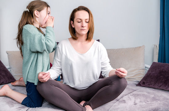 Calm mindful single mom doing yoga at home while active noisy kid daughter screaming in her ear, mother meditating for stress relief and emotional balance relaxing in living room with child daughter