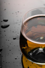 Full glass of nice gold colour fresh beer with air bubbles and white foam on black reflective surface with sprayed water drops on it