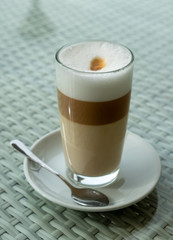 Glass with latte coffee on a saucer with a spoon. A table in a street cafe. White borders of milk and brown edges of coffee are visible in a glass. Clear day. The city of Tbilisi, Georgia.