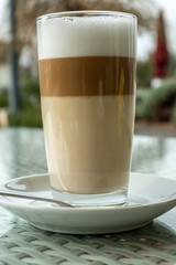 Glass with latte coffee on a saucer with a spoon. A table in a street cafe. White borders of milk and brown edges of coffee are visible in a glass. Clear day. The city of Tbilisi, Georgia.