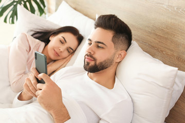 Young man using mobile phone while his wife sleeping in bed