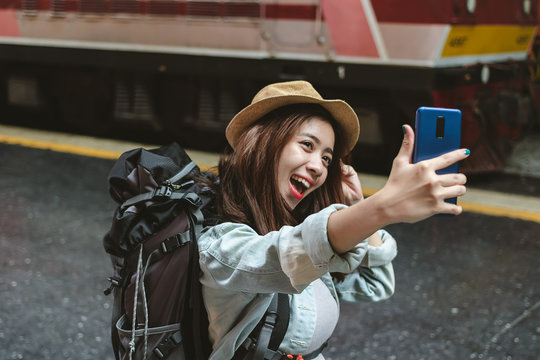 Cheerful young Asian woman traveler with backpack taking a photo or selfie in train station. Travel lifestyle concept.