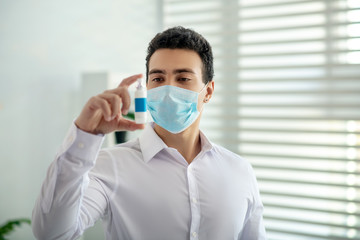 Dark-haired man in a protective mask holding a disinfector in his hand an looking serious