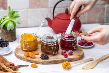 Female hands preparing a raw vegan raspberry jam on organic brown bread on a white wooden kitchen table. Real scene with organic fruit jam.