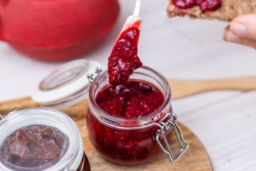 Raspberry jam in a glass jar with female hand grabbing with white spoon on a wooden plate.