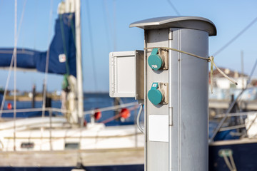 An aluminum power connection box in the marina of the small German port of Scharprode on the island of Rügen. In the background is a sailboat on a jetty.