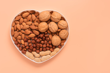 A variety of nuts in a bowl a heart shape on a pink pastel background. Almonds, walnuts, hazelnuts, peanuts. Top view, flat lay, place for text.