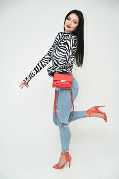 Model glamorous girl in blue jeans and a striped blouse stands right with a red bag on a white background. Vertical photo of a fashionable caucasian brunette. Side view.