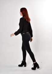 Portrait of a pretty girl with red hair wearing black jeans, boots and a blouse.  full length standing pose on a studio background, with back to the camera.
