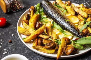 Baked mackerel with lemon, baked potatoes and mushrooms on a plate on black stone background.