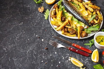 Baked mackerel with lemon, baked potatoes and mushrooms on a plate on black stone background.