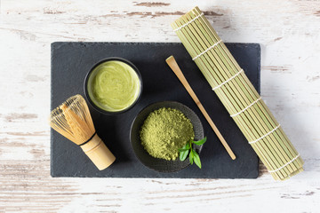 Fototapeta na wymiar Green matcha tea drink and tea accessories on black board on white wooden table top view. Japanese tea ceremony concept.