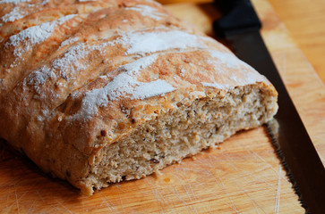 A loaf of home-baked sourdough bread on a wooden chopping board on a wooden table with a large black-handled knife in natural lighting
