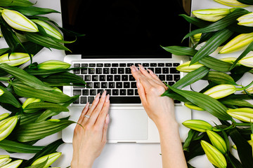 A woman printing on a laptop keyboard which surrounded with green flowers. Trendy composition in...