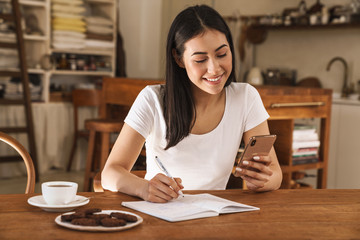 Image of smiling woman making notes in planner and using mobile phone