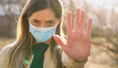 Young woman wearing blue disposable single use virus mouth nose mask, hold hand front of her as stop gesture, blurred sunset lit trees in background. Coronavirus covid-19 outbreak prevention concept