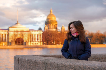 A brunette girl with long hair in a jacket and scarf stands on the embankment of St. Petersburg at sunset with a view of St. Isaac's Cathedral and the Golden buildings