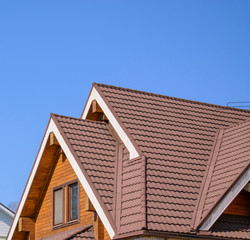 Gable triangular roof with a large pediment. Glued beams with grooves, attic window, brown tiles. Blue sky background.