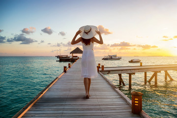 A beautiful tourist woman in white dress on summer vacations walks over a wooden pier into the tropical sunset on the Maldives islands - obrazy, fototapety, plakaty