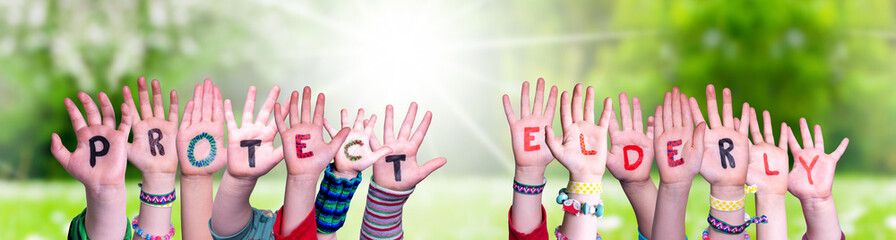 Kids Hands Holding Colorful English Word Protect Elderly. Sunny Green Grass Meadow As Background