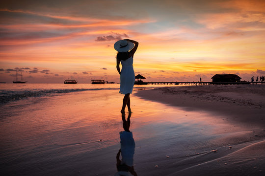 Eine Frau mit Hut steht am tropischen Strand bei Sonnenuntergang und genie&szlig;t die Atmosph&auml;re der Malediven