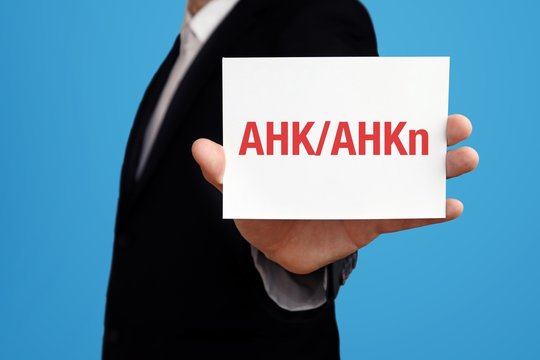 AHK/AHKn. Gesch&auml;ftsmann im Anzug h&auml;lt Karte in die Kamera. Der Begriff AHK/AHKn steht im Schild. Symbol f&uuml;r Business, Finanzen, Statistik, Analyse, Wirtschaft