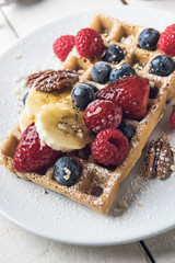 Waffle topped with banana slices, oat, pecans and berries served on a white plate over a white rustic wooden table. Closeup.